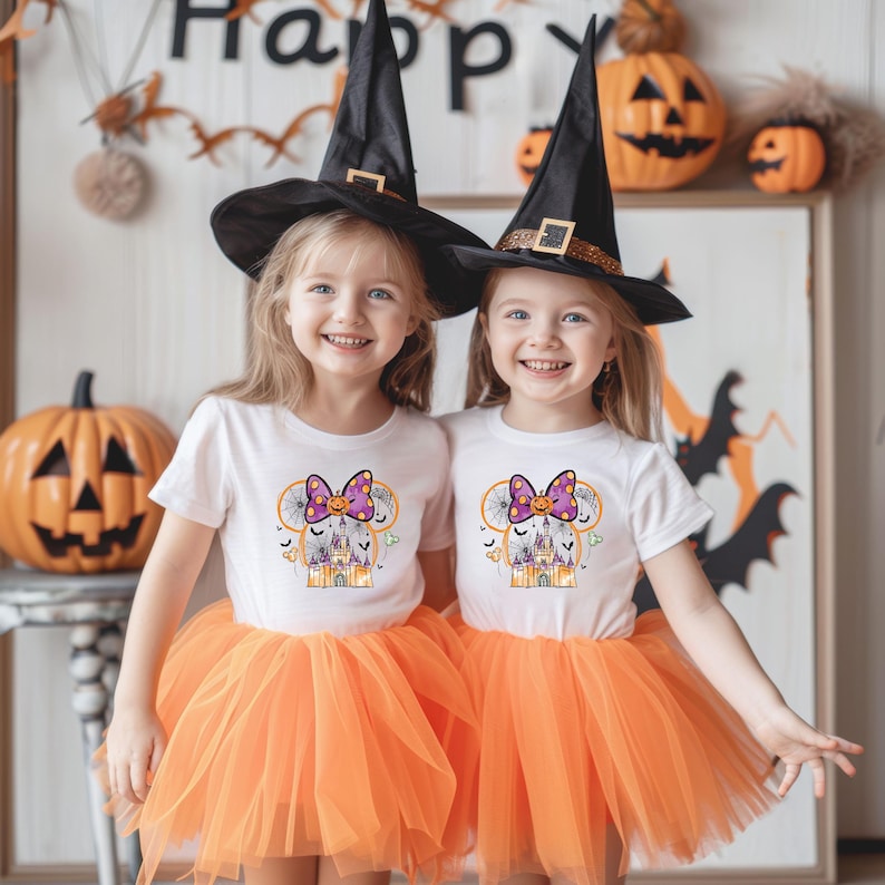 May include: Two young girls wearing matching Halloween costumes. Each girl wears a white t-shirt with a graphic, an orange tutu, and a black witch hat. The graphic on the shirt features a purple bow, a castle, and a Mickey Mouse-inspired design.