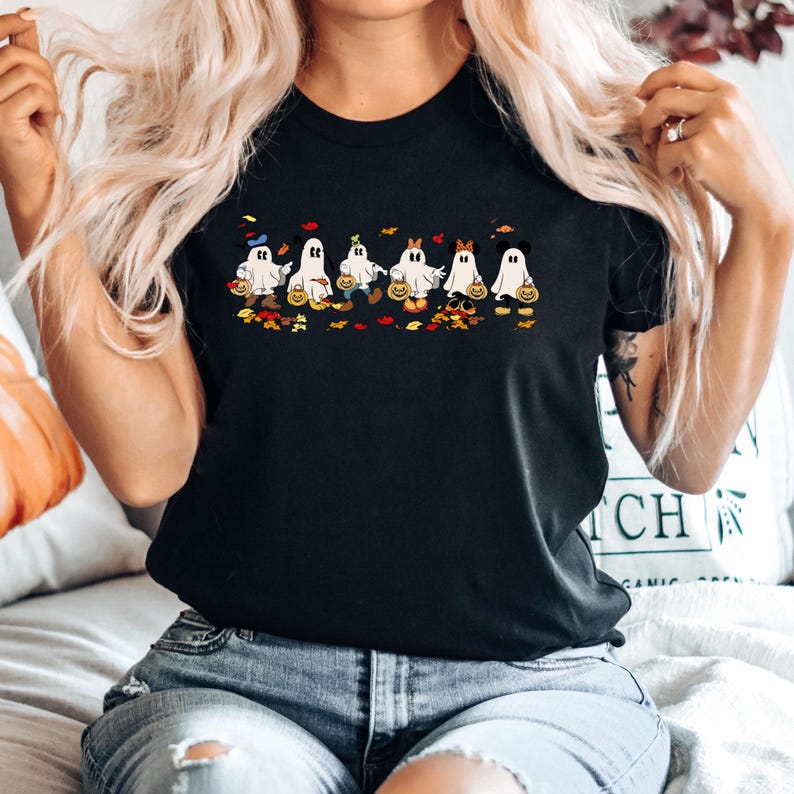 May include: Black t-shirt featuring a Halloween-themed design. The graphic shows a row of cartoon ghosts dressed as popular characters, each holding a pumpkin bucket. Colorful leaves and pumpkins are scattered below the ghosts.