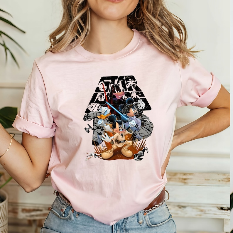 May include: Pink t-shirt with a black and white graphic of Mickey Mouse, Donald Duck, and Minnie Mouse dressed as Star Wars characters. The graphic includes the text "Star Wars" in a black triangle.