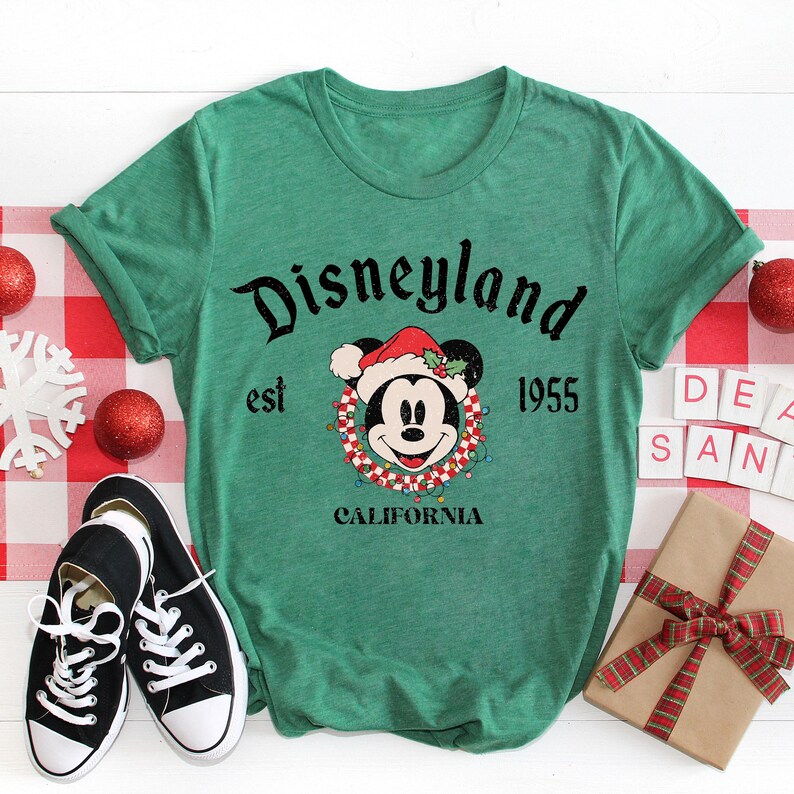 Puede incluir: Camiseta verde con un logotipo vintage de Disneyland con Mickey Mouse con un gorro de Pap&aacute; Noel. El texto dice "Disneyland est 1955 California".