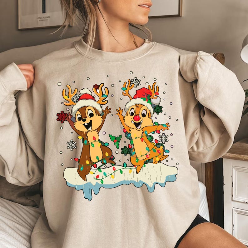 May include: A beige sweatshirt with a Christmas-themed design featuring two cartoon chipmunks wearing Santa hats and surrounded by Christmas lights. The chipmunks are standing on a snowy surface.