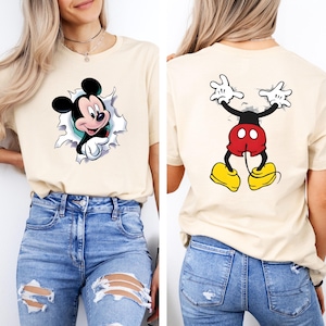 Puede incluir: Camiseta beige con un gr&aacute;fico de Mickey Mouse. La parte delantera de la camiseta muestra a Mickey rompiendo un dise&ntilde;o rasgado, mientras que la parte trasera muestra la parte trasera de Mickey. La camiseta se usa con jeans azules.