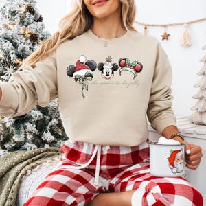 May include: Beige sweatshirt featuring Mickey Mouse ears with holiday decorations. The design includes a Santa hat, a festive cake, and holly. The text "the season to be jolly" is printed below the ears. The person is wearing red and white plaid pajama pants.