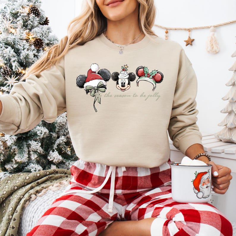 May include: Beige sweatshirt featuring Mickey Mouse ears with holiday decorations. The design includes a Santa hat, a festive cake, and holly. The text "the season to be jolly" is printed below the ears. The person is wearing red and white plaid pajama pants.