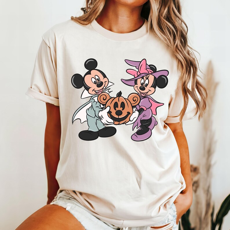 May include: A white t-shirt with a graphic of Mickey Mouse and Minnie Mouse dressed in Halloween costumes. Mickey is dressed as a vampire and Minnie is dressed as a witch. They are holding a jack-o'-lantern.