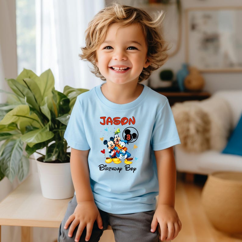 May include: A light blue t-shirt featuring Mickey Mouse, Donald Duck, and Goofy. The shirt has the name "Jason" in red, the number "3" in a Mickey Mouse head, and the words "Birthday Boy" in black.