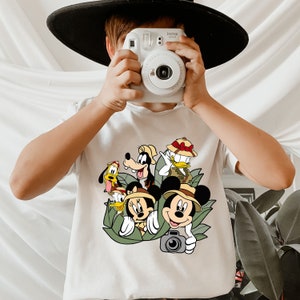 May include: White t-shirt featuring a graphic print of Mickey Mouse, Minnie Mouse, Goofy, Pluto and Donald Duck in a jungle setting. The characters are wearing safari hats and holding cameras. The text "Instax" is visible on a camera in the image.