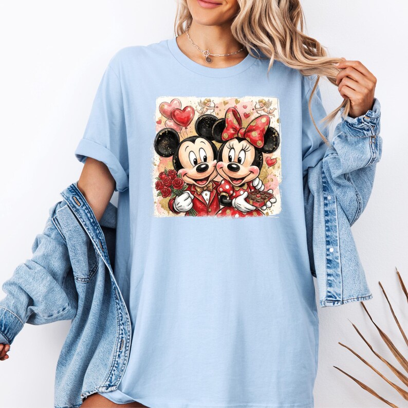 May include: Light blue t-shirt featuring a graphic of Mickey and Minnie Mouse in Valentine's Day attire. Mickey is holding roses, and Minnie holds a treat. The image includes heart-shaped balloons and a decorative background.