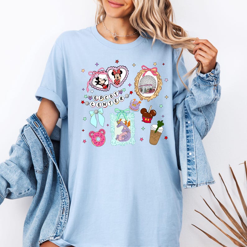 May include: Light blue t-shirt with a graphic design featuring various Disney characters and icons from Epcot Center. The design includes Mickey Mouse, Minnie Mouse, Spaceship Earth, a donut, a dragon, and a coffee cup.