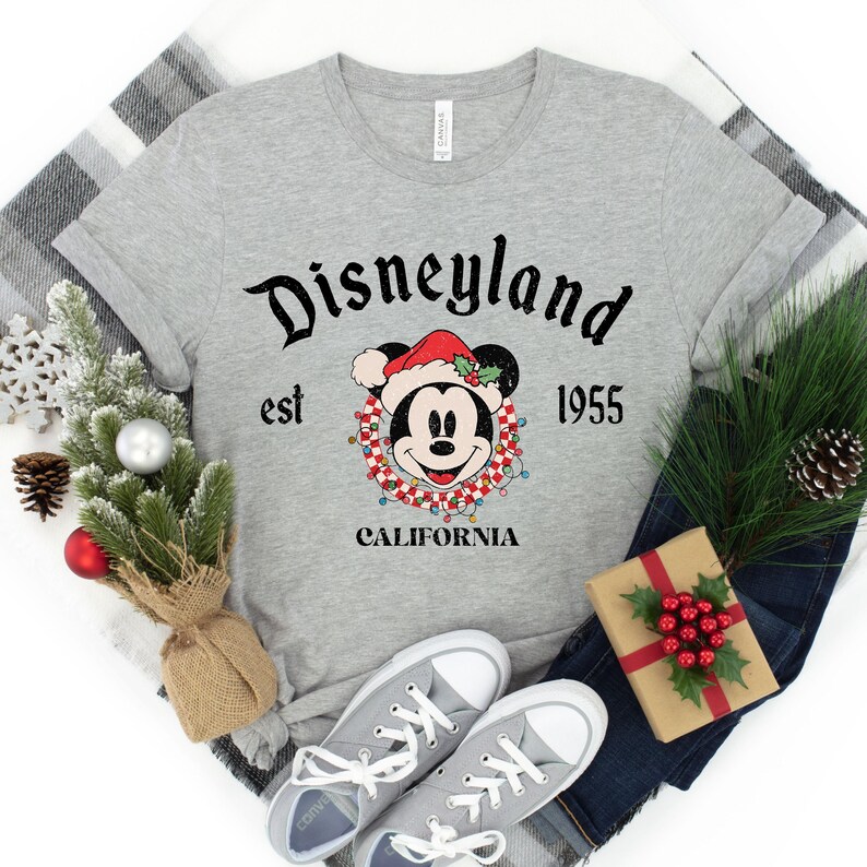 Puede incluir: Camiseta gris con un logotipo vintage de Disneyland con Mickey Mouse con un gorro de Pap&aacute; Noel y una corona roja y blanca. El texto dice "Disneyland est 1955 California".
