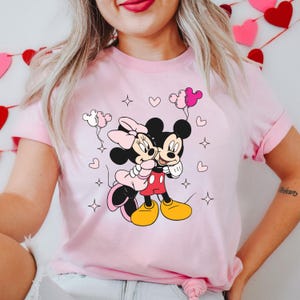 May include: Pink t-shirt with a graphic of Mickey Mouse and Minnie Mouse holding each other. The graphic is surrounded by hearts and stars.
