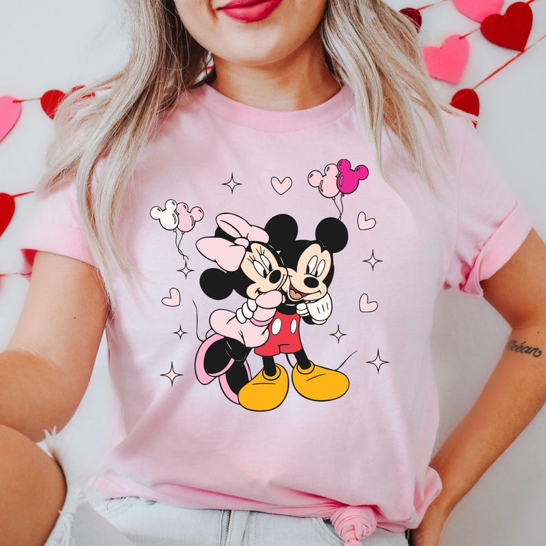 May include: Pink t-shirt with a graphic of Mickey Mouse and Minnie Mouse holding each other. The graphic is surrounded by hearts and stars.