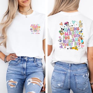 May include: White t-shirt with a colorful graphic on the front featuring a Minnie Mouse design and the text "Mrs. White." The back of the shirt has a graphic with the text "MAKING MAGIC FOR TINY HUMANS EVERY DAY" and cartoon characters.
