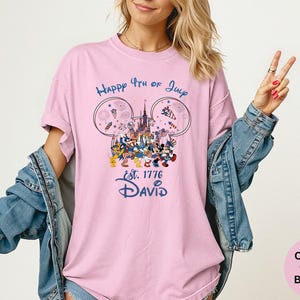 May include: A light pink t-shirt with a graphic featuring Disney characters celebrating the 4th of July. The design includes fireworks, a castle, and the text "Happy 4th of July Est. 1776 David" in blue. The shirt is a comfort colors blossom.