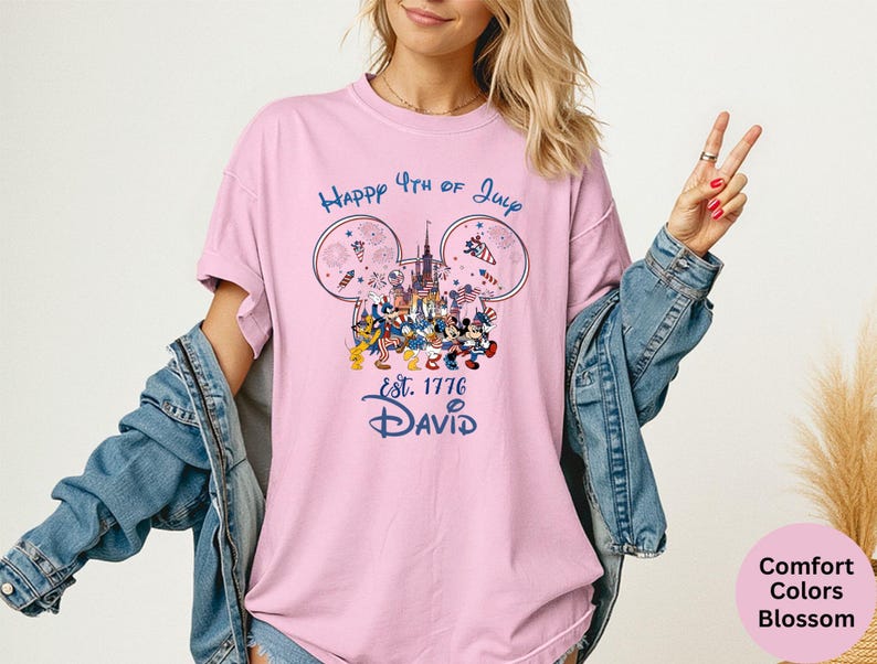 May include: A light pink t-shirt with a graphic featuring Disney characters celebrating the 4th of July. The design includes fireworks, a castle, and the text "Happy 4th of July Est. 1776 David" in blue. The shirt is a comfort colors blossom.
