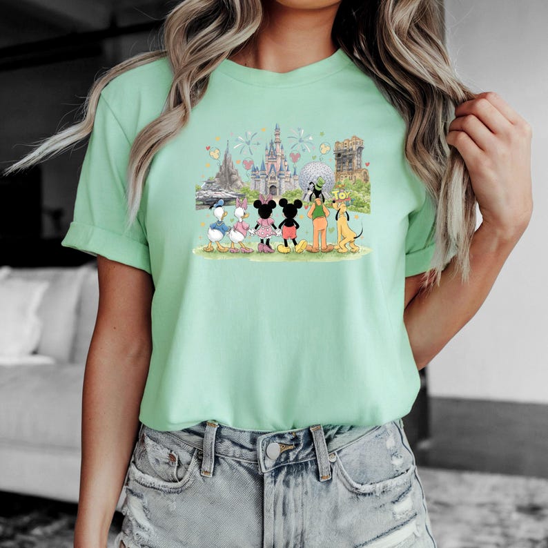 May include: A light green t-shirt featuring a graphic of Disney characters including Donald Duck, Daisy Duck, Mickey Mouse, Minnie Mouse, Goofy, and Pluto. The graphic also includes images of Disney landmarks such as Cinderella's castle.