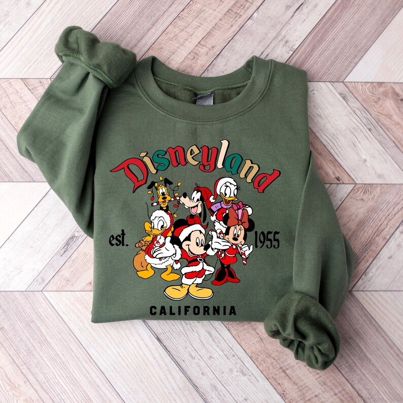 May include: Green sweatshirt with a Christmas-themed Disneyland graphic featuring Mickey Mouse, Minnie Mouse, Donald Duck, Goofy, and Pluto. The graphic includes the text "Disneyland est. 1955 California".