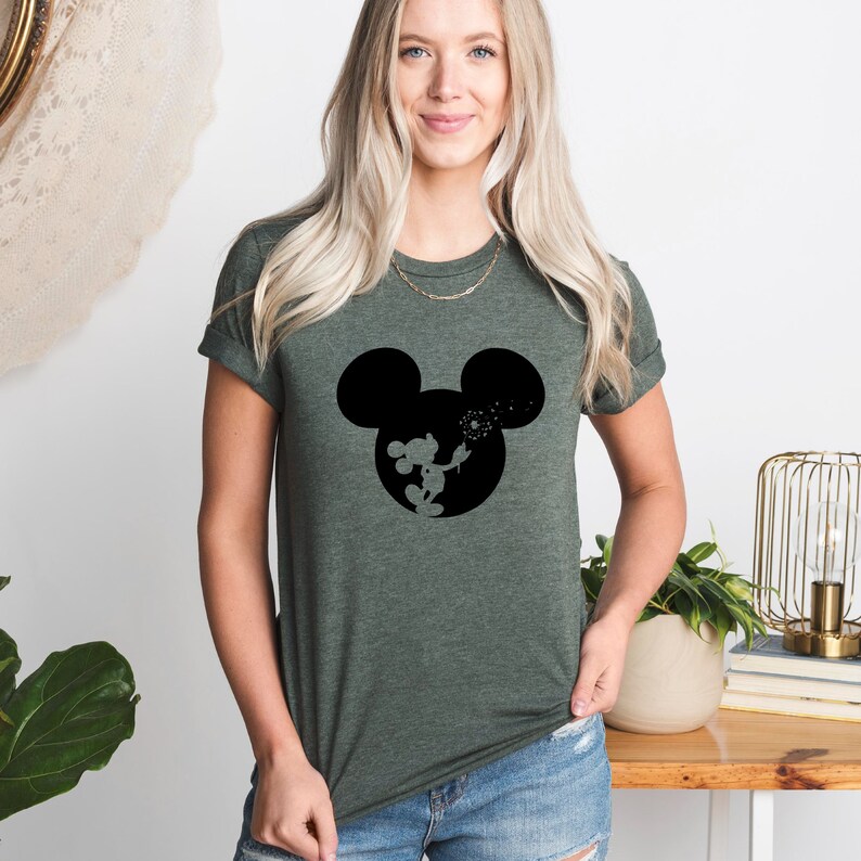 May include: A green t-shirt with a black silhouette of Mickey Mouse blowing on a dandelion. The silhouette is in the center of the shirt.