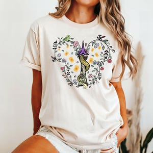 May include: A white t-shirt with a floral design featuring a purple tower and a long braid of hair. The design is in the shape of a Mickey Mouse head.