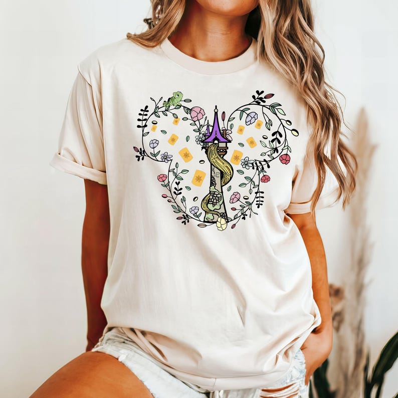May include: A white t-shirt with a floral design featuring a purple tower and a long braid of hair. The design is in the shape of a Mickey Mouse head.