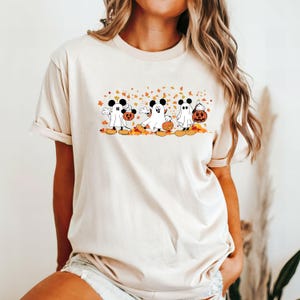 May include: A cream colored t-shirt with a graphic of three Mickey Mouse characters dressed as ghosts holding pumpkins. The characters are walking in a line with autumn leaves falling around them.