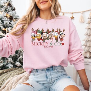 K&ouml;nnte beinhalten: Hellrosa Sweatshirt mit einem weihnachtlichen Motiv von Mickey Mouse und seinen Freunden. Die Charaktere tragen Rentiergeweihe und Schals. Der Text "Mickey & Co." und "Est. 1928" ist rot gedruckt.