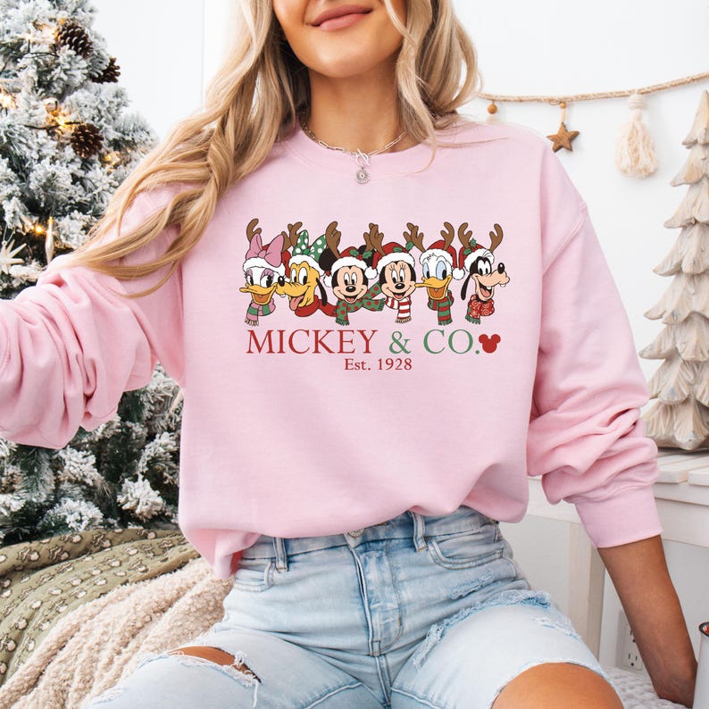 K&ouml;nnte beinhalten: Hellrosa Sweatshirt mit einem weihnachtlichen Motiv von Mickey Mouse und seinen Freunden. Die Charaktere tragen Rentiergeweihe und Schals. Der Text "Mickey & Co." und "Est. 1928" ist rot gedruckt.