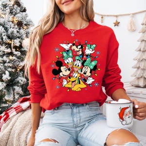 K&ouml;nnte beinhalten: Ein rotes Sweatshirt mit einem festlichen Design mit den Disney-Figuren Mickey Mouse, Minnie Mouse, Donald Duck, Daisy Duck, Goofy und Pluto, umgeben von Lichterketten und Schneeflocken. Das Sweatshirt wird mit hellblauen Jeans getragen.