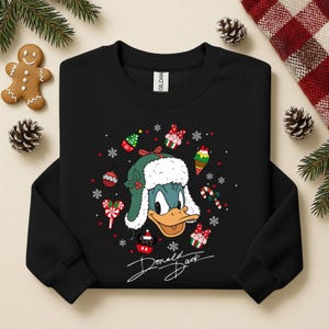 May include: Black sweatshirt featuring a graphic of Donald Duck wearing a green and white winter hat. The design includes Christmas-themed elements like ornaments, snowflakes, and candy canes. The words "Donald Duck" are written in a cursive font.