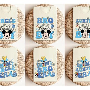 Op de afbeelding: Cr&egrave;mekleurige T-shirts met blauwe tekst en Mickey Mouse-afbeeldingen. De shirts tonen zinnen als "Uncle Birthday Boy", "Bro Birthday Boy" en "In My Uncle Era". Elk gevouwen T-shirt ligt op een geweven mat.