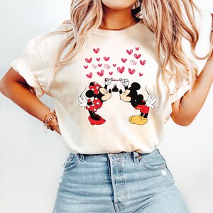 Puede incluir: Camiseta color crema con un gr&aacute;fico de Mickey y Minnie Mouse bes&aacute;ndose, rodeados de corazones rojos. El dise&ntilde;o incluye la palabra "Love" y una silueta de castillo. Los personajes visten sus trajes cl&aacute;sicos.