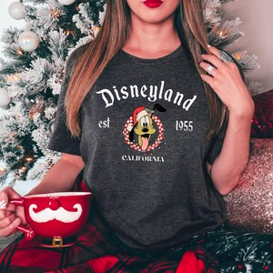 May include: A gray t-shirt with a white graphic of Pluto wearing a Santa hat and a wreath. The graphic also includes the text "Disneyland est 1955 California".