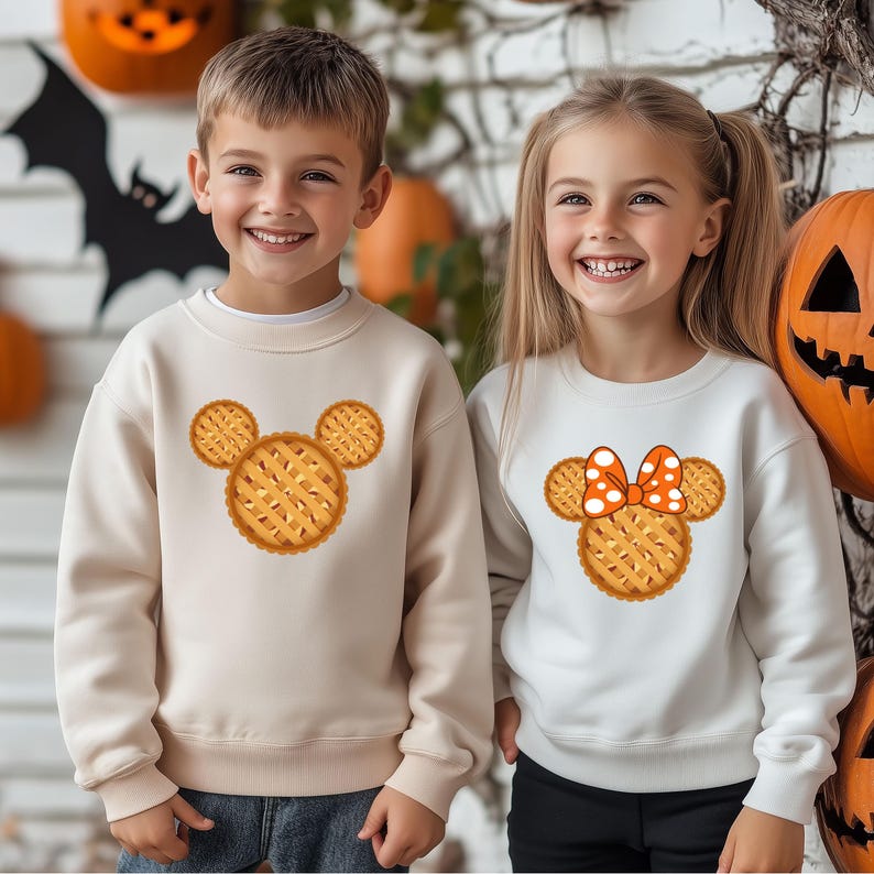 K&ouml;nnte beinhalten: Zwei Kinder tragen cremefarbene Sweatshirts mit einem Kuchen-Design in Form einer Maus. Ein Sweatshirt hat eine Schleife mit orangefarbenen Punkten. Halloween-Dekorationen sind im Hintergrund zu sehen.