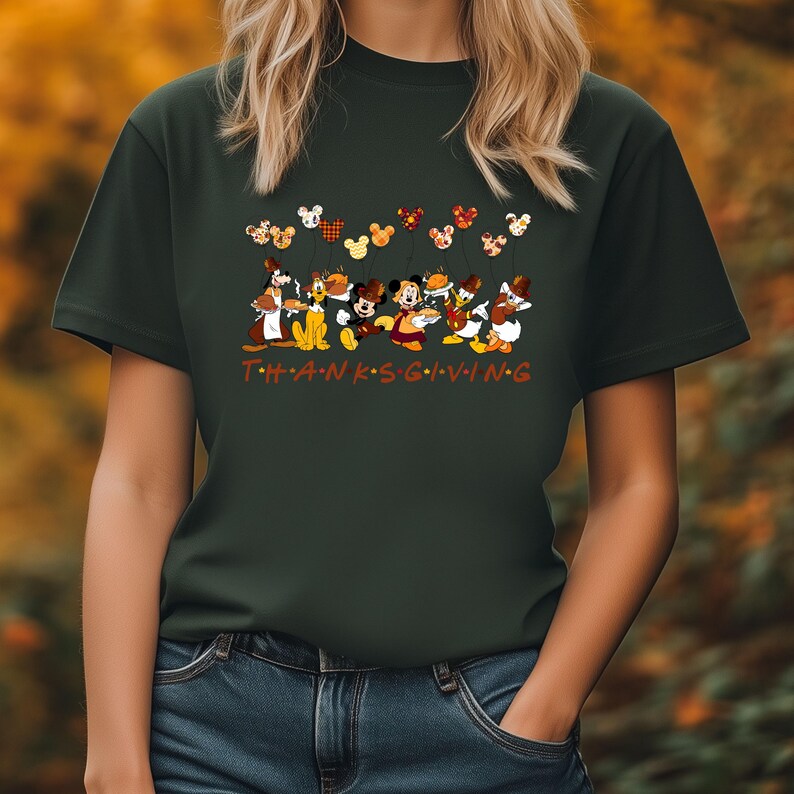 Pu&ograve; includere: T-shirt verde scuro con un design a tema Ringraziamento. La grafica include personaggi Disney come Topolino, Paperino e Pippo, con la scritta "THANKSGIVING" in arancione. Il design presenta anche forme di testa di Topolino.