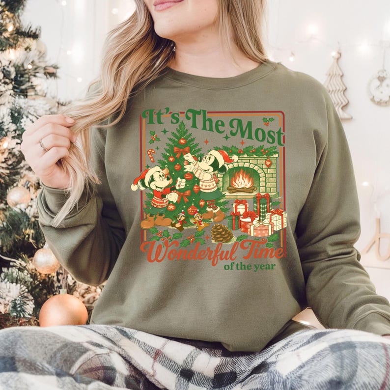 May include: Khaki green sweatshirt with a festive Christmas graphic. The design shows Mickey and Minnie Mouse by a decorated tree, a fireplace, and presents. The text reads "It's The Most Wonderful Time of the Year."