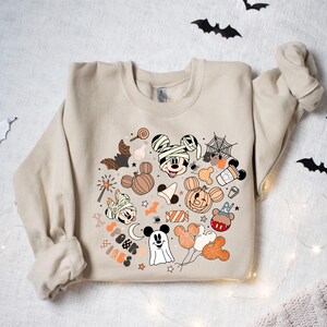 May include: A beige sweatshirt with a Halloween-themed design featuring Mickey Mouse and friends. The design includes Mickey Mouse dressed as a mummy, a ghost, a pumpkin, and other Halloween icons. The text "Spook Vibes" is also included in the design.