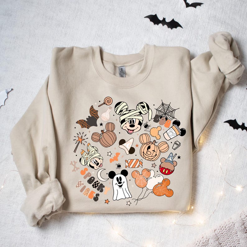 May include: A beige sweatshirt with a Halloween-themed design featuring Mickey Mouse and friends. The design includes Mickey Mouse dressed as a mummy, a ghost, a pumpkin, and other Halloween icons. The text "Spook Vibes" is also included in the design.