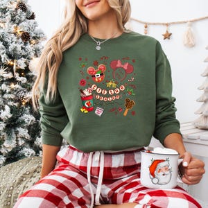 May include: Green sweatshirt with a festive graphic that includes a Mickey Mouse cookie, Minnie Mouse ears, and the phrase "Tis the Season." The sweatshirt is paired with red and white plaid pajama pants. A white mug with a Santa Claus design is held in the hand.