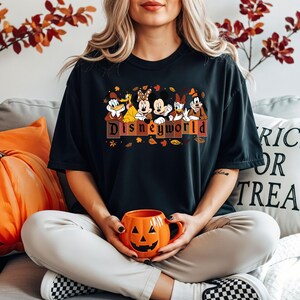 May include: Black t-shirt featuring Disney characters with the text "Disneyworld" in brown and orange. The shirt is worn with white pants and black and white checkered shoes. An orange pumpkin mug is held in the hands.