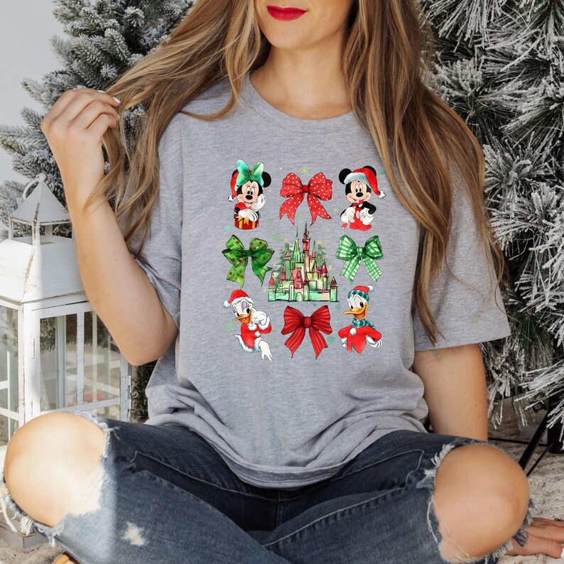 May include: A gray t-shirt with a Christmas-themed design featuring Mickey Mouse, Minnie Mouse, Donald Duck, and the Disney castle. The design includes red and green bows and a watercolor effect.