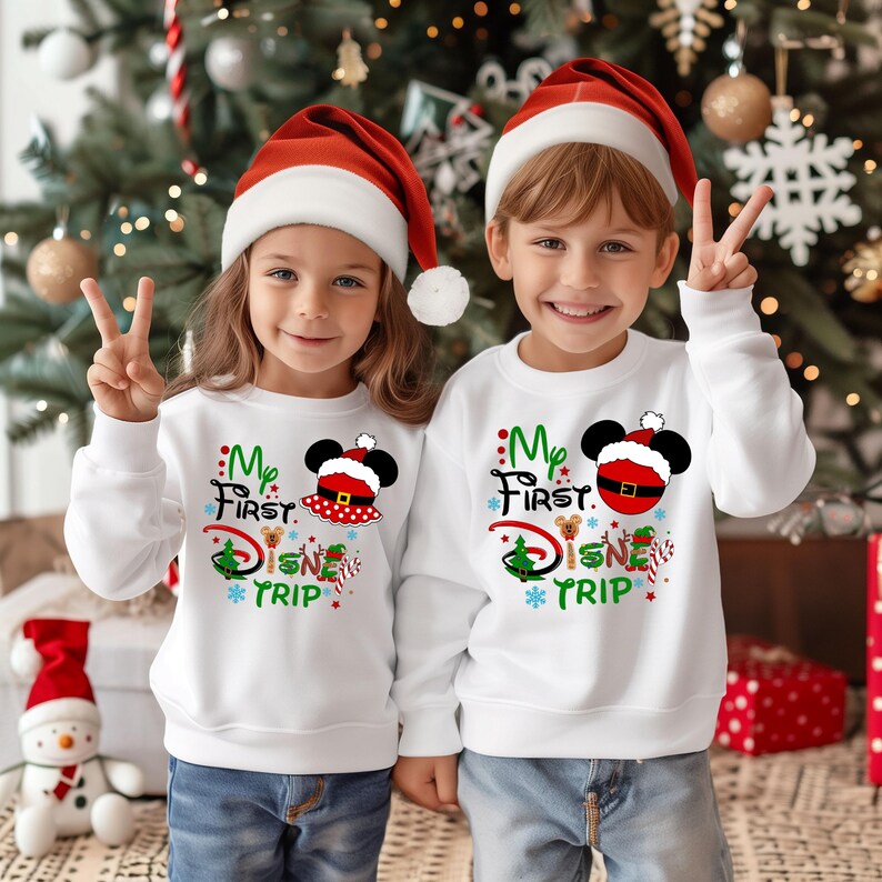 May include: Two children wearing white sweatshirts with a Christmas Mickey Mouse design that says "My First Disney Trip". They are wearing red Santa hats and smiling.