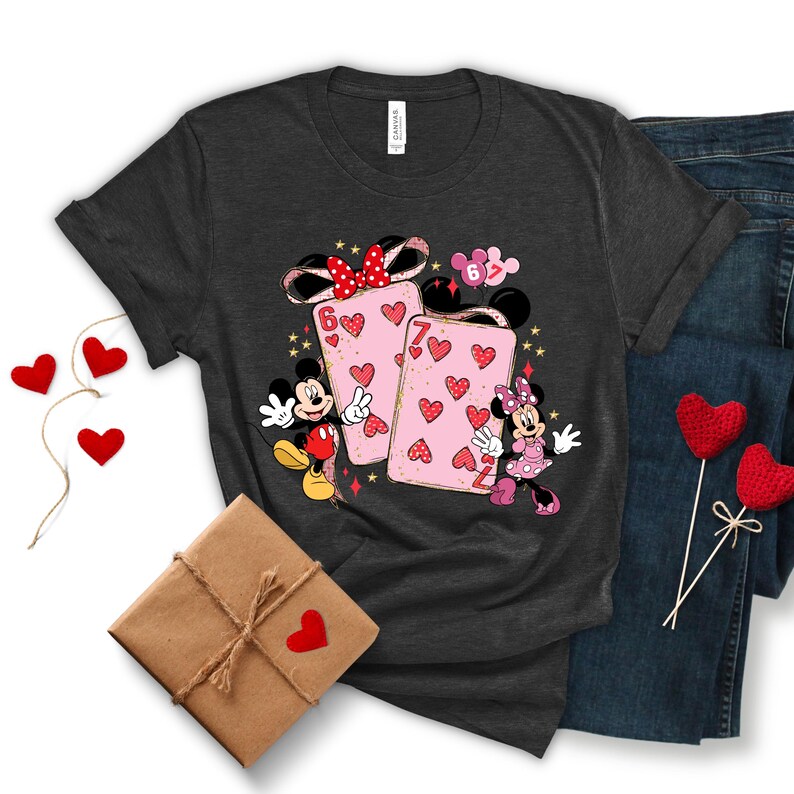 May include: Dark gray t-shirt featuring Mickey and Minnie Mouse with playing cards decorated with hearts. The cards have the numbers 6 and 7. The shirt is paired with blue jeans, a wrapped gift, and heart-shaped decorations.