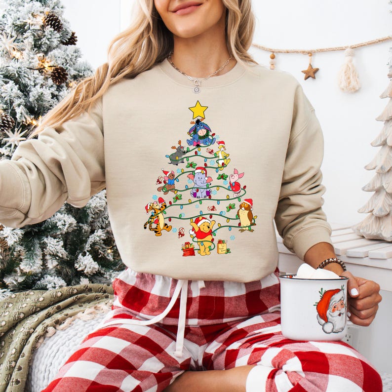 Pu&ograve; includere: Felpa beige con un design ad albero di Natale realizzato con personaggi dei cartoni animati, tra cui Winnie the Pooh e amici. L'albero &egrave; decorato con luci e una stella. La persona indossa pantaloni del pigiama a quadri rossi e bianchi e tiene in mano una tazza con un disegno di Babbo Natale.