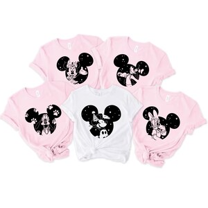 May include: Five short-sleeved t-shirts in pink and white. Each shirt features a black Mickey Mouse silhouette with different character designs. The designs include Minnie Mouse, Goofy, Mickey Mouse, and Donald Duck.