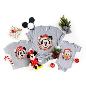 May include: A set of three grey t-shirts featuring a Christmas-themed design with Mickey Mouse, Minnie Mouse, and Donald Duck. The shirts are decorated with a red and green wreath, holly, and lights.