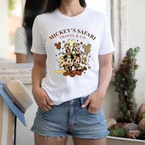 K&ouml;nnte beinhalten: Wei&szlig;es T-Shirt mit einem Aufdruck von Mickey Mouse und seinen Freunden in Safari-Kleidung. Der Text "Mickey's Safari Travel & Co" ist auf dem Shirt aufgedruckt.