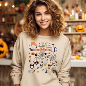 May include: A beige sweatshirt with a Halloween-themed design featuring Mickey Mouse, Donald Duck, Goofy, and other Disney characters. The design includes a spiderweb, pumpkins, and the text "World Walt Disney".