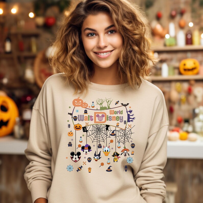 May include: A beige sweatshirt with a Halloween-themed design featuring Mickey Mouse, Donald Duck, Goofy, and other Disney characters. The design includes a spiderweb, pumpkins, and the text "World Walt Disney".