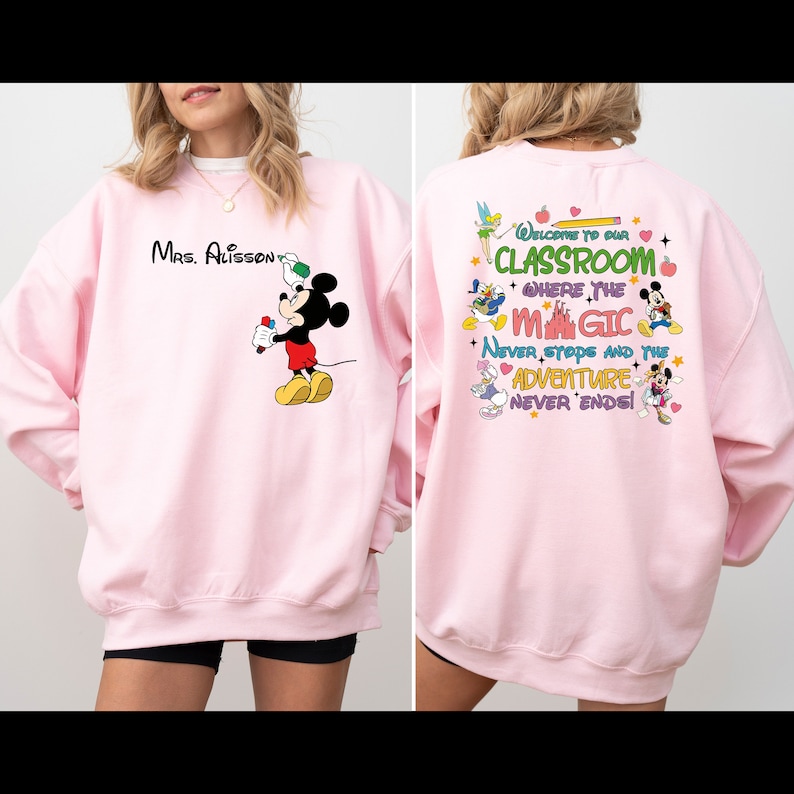 May include: Pink sweatshirt with a Mickey Mouse design on the front and a Disney-themed design on the back. The back design includes the text 'Welcome to our Classroom where the magic never stops and the adventure never ends!'.