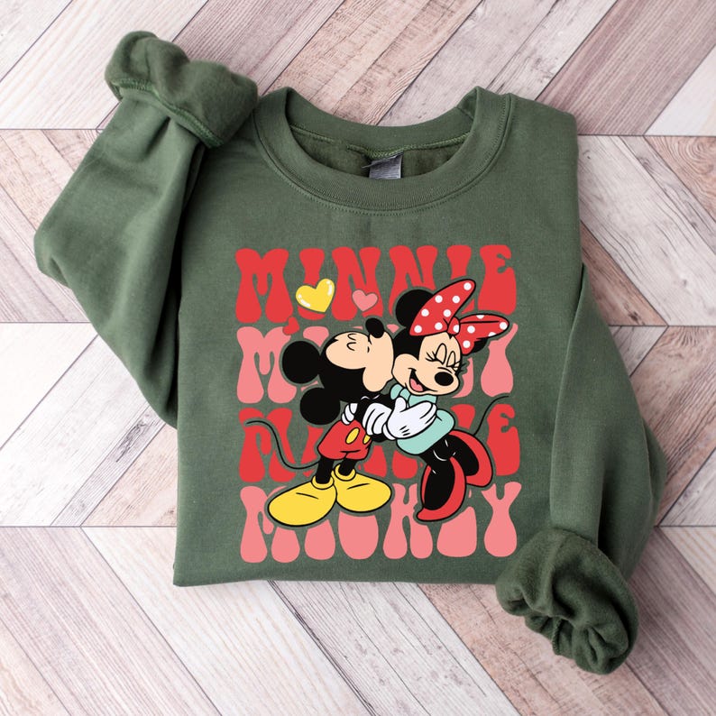 K&ouml;nnte beinhalten: Olivgr&uuml;nes Sweatshirt mit einer Grafik von Mickey und Minnie Mouse, die sich umarmen. Der Text "MINNIE MICKEY" ist in roter und rosa Retro-Schrift, mit einem gelben Herz. Die &Auml;rmel sind ger&uuml;scht.